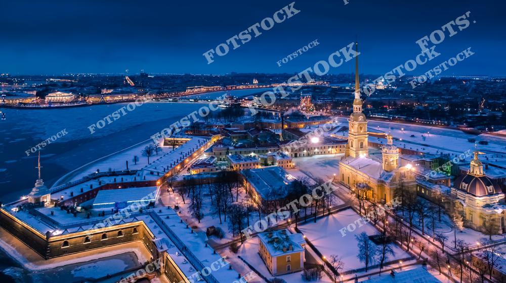 На фотографии изображена Петропавловская крепость, ярко освещенная в зимнюю ночь. Собор святых Петра и Павла с его высоким шпилем доминирует в центре композиции. Заснеженные территории крепости и замерзшая река Нева создают атмосферу покоя и величия. Вдали видны огни других достопримечательностей Санкт-Петербурга, добавляющие шарма ночному пейзажу.